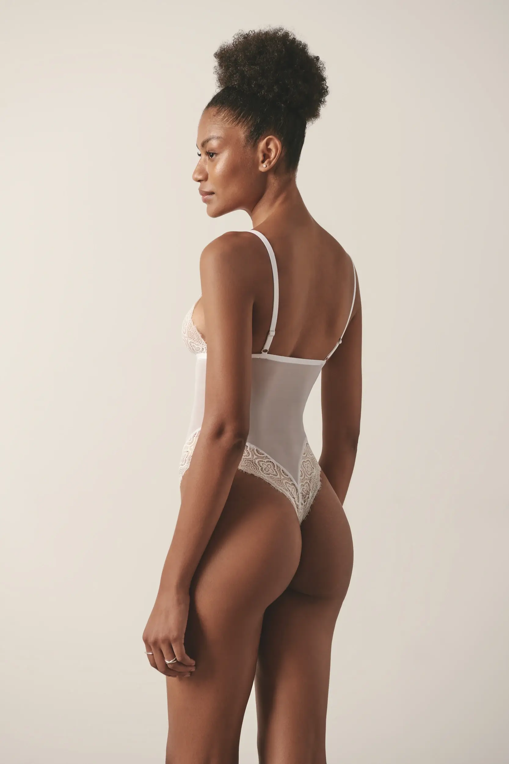 Back view of a model wearing a white mesh and lace lingerie bodysuit, posing in a studio