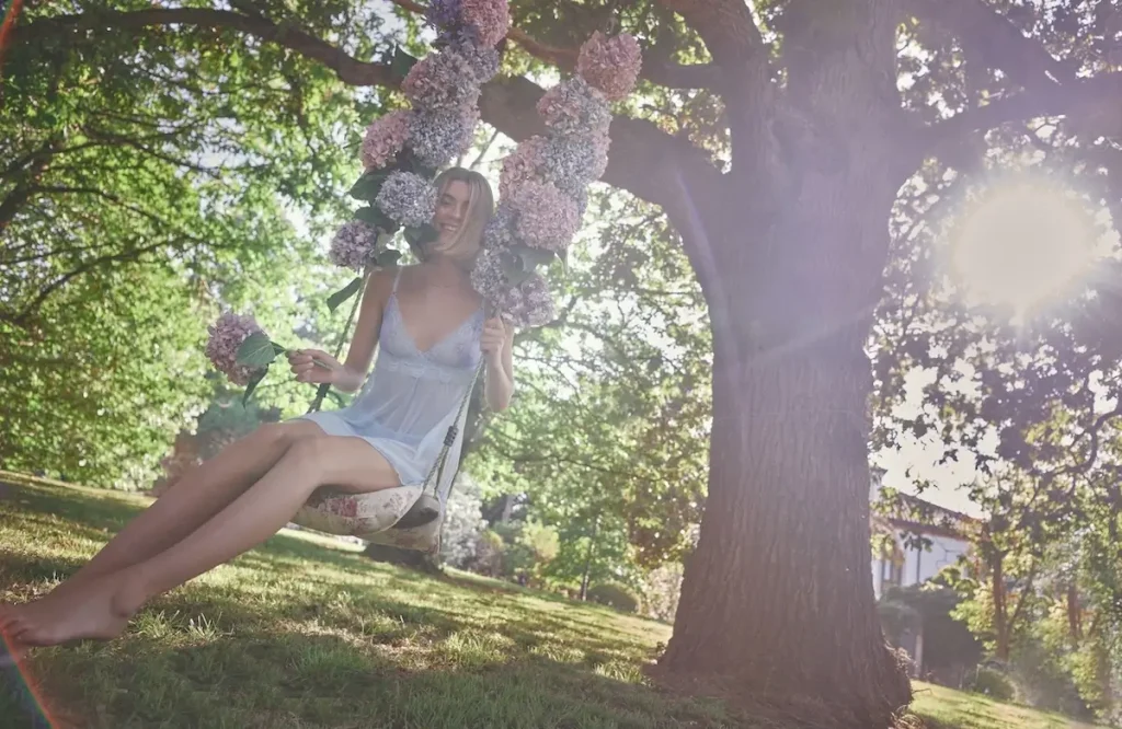 A woman wearing a lavender lingerie baby doll sits on a swing hanging from a tree in a garden, laughing and surrounded by flowers